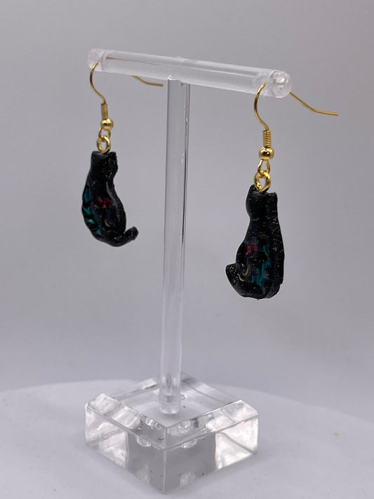Two black cat dangle earrings with colorful glitter accents hanging from gold hooks on a clear plastic display stand.